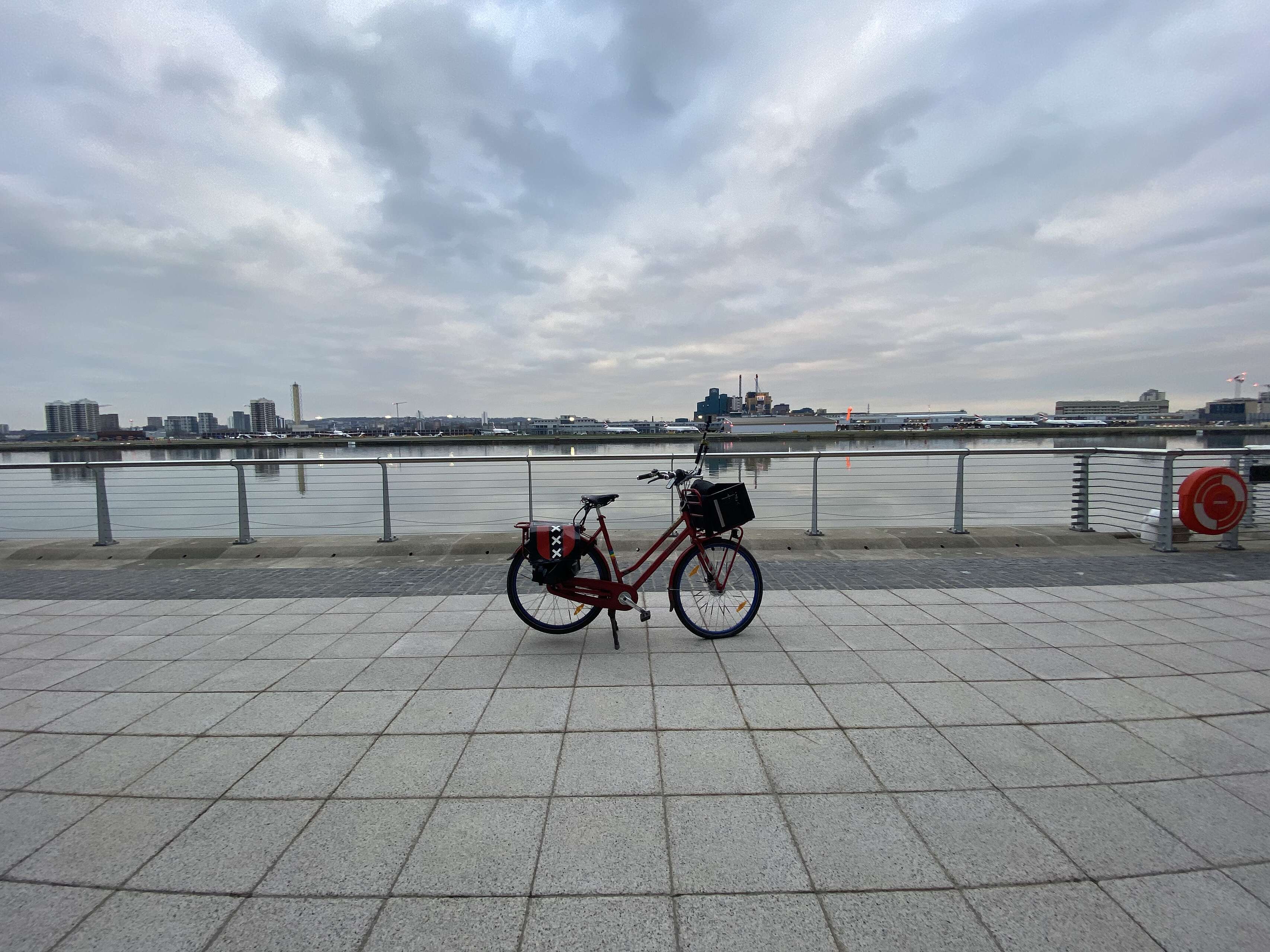Red Dutch bicycle on its kickstand on a paved area in front of a dock with London City Airport in the background across the dock.jpeg